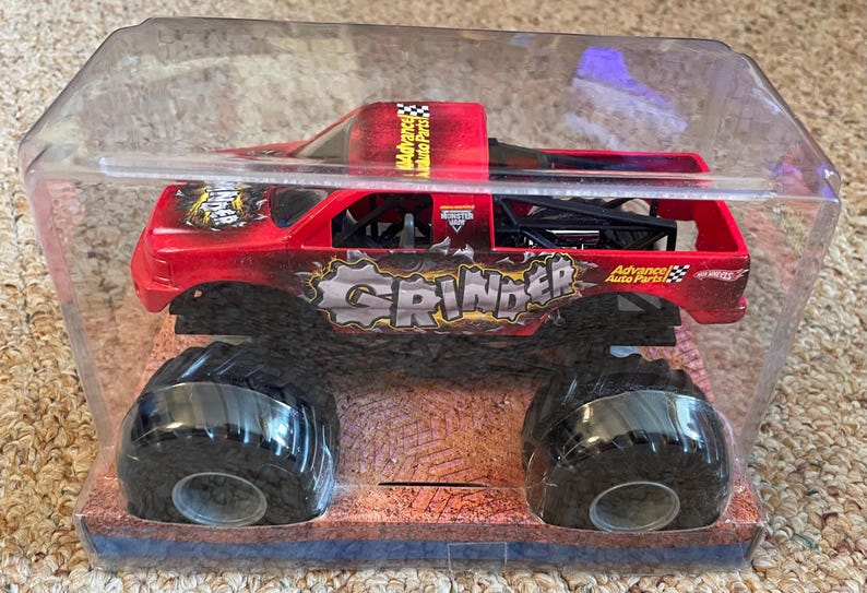 May include: A red and black toy monster truck named "Grinder" in a clear plastic package. The truck has large black tires and the words "Advance Auto Parts" are visible. The truck is a toy vehicle.