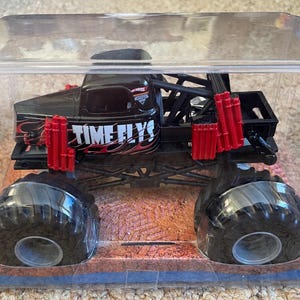 May include: Black monster truck toy with large black tires, red accents, and the text "TIME FLYS" on the side. The toy is encased in a clear plastic display case, sitting on a brown and blue base.
