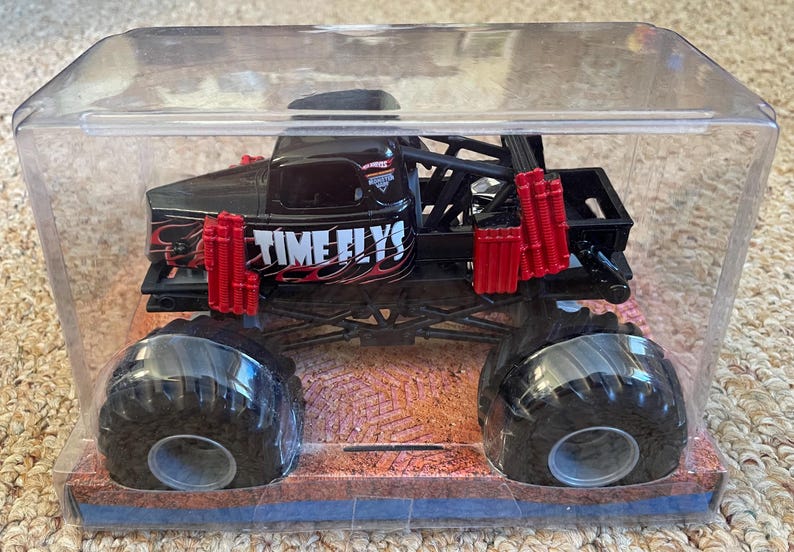 May include: Black monster truck toy with large black tires, red accents, and the text "TIME FLYS" on the side. The toy is encased in a clear plastic display case, sitting on a brown and blue base.