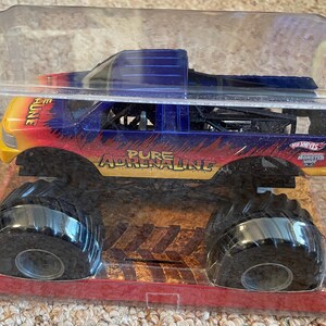 Może przedstawiać: Zabawka monster truck Hot Wheels w przezroczystym plastikowym opakowaniu. Ciężar&oacute;wka ma niebieski, ż&oacute;łty i czerwony schemat malowania z napisem "PURE ADRENALINE" z boku. Ciężar&oacute;wka ma duże czarne opony.