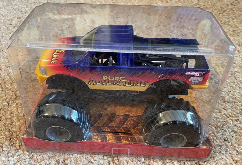 Może przedstawiać: Zabawka monster truck Hot Wheels w przezroczystym plastikowym opakowaniu. Ciężar&oacute;wka ma niebieski, ż&oacute;łty i czerwony schemat malowania z napisem "PURE ADRENALINE" z boku. Ciężar&oacute;wka ma duże czarne opony.