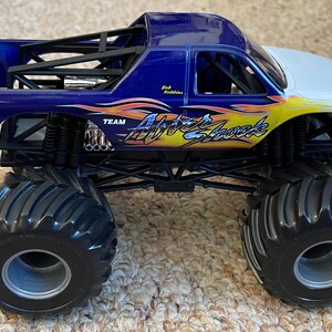 May include: A blue, white, and yellow Hot Wheels monster truck toy. The truck features the words "After Shock" and "Team" on the side. The truck has large black tires and a roll cage.
