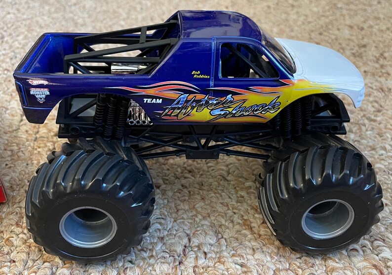 May include: A blue, white, and yellow Hot Wheels monster truck toy. The truck features the words "After Shock" and "Team" on the side. The truck has large black tires and a roll cage.