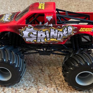 May include: A red toy monster truck with black wheels and a black roll cage. The truck has the word "GRINDER" in a distressed font on the side, along with "Advance Auto Parts" branding. The tires are large and black.