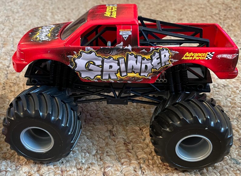 May include: A red toy monster truck with black wheels and a black roll cage. The truck has the word "GRINDER" in a distressed font on the side, along with "Advance Auto Parts" branding. The tires are large and black.