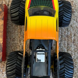 May include: A toy monster truck with a gradient yellow to orange body, black tires, and a green grid design on the roof. The truck has a detailed engine and chassis.
