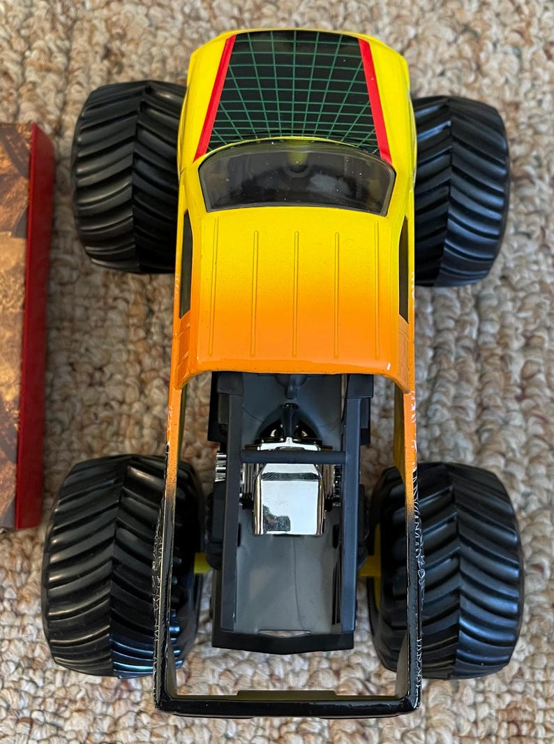 May include: A toy monster truck with a gradient yellow to orange body, black tires, and a green grid design on the roof. The truck has a detailed engine and chassis.