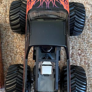 May include: A black monster truck toy with large black tires. The top of the truck is orange with a flame design. The truck's frame is black, and the engine is visible. The toy is designed for children.