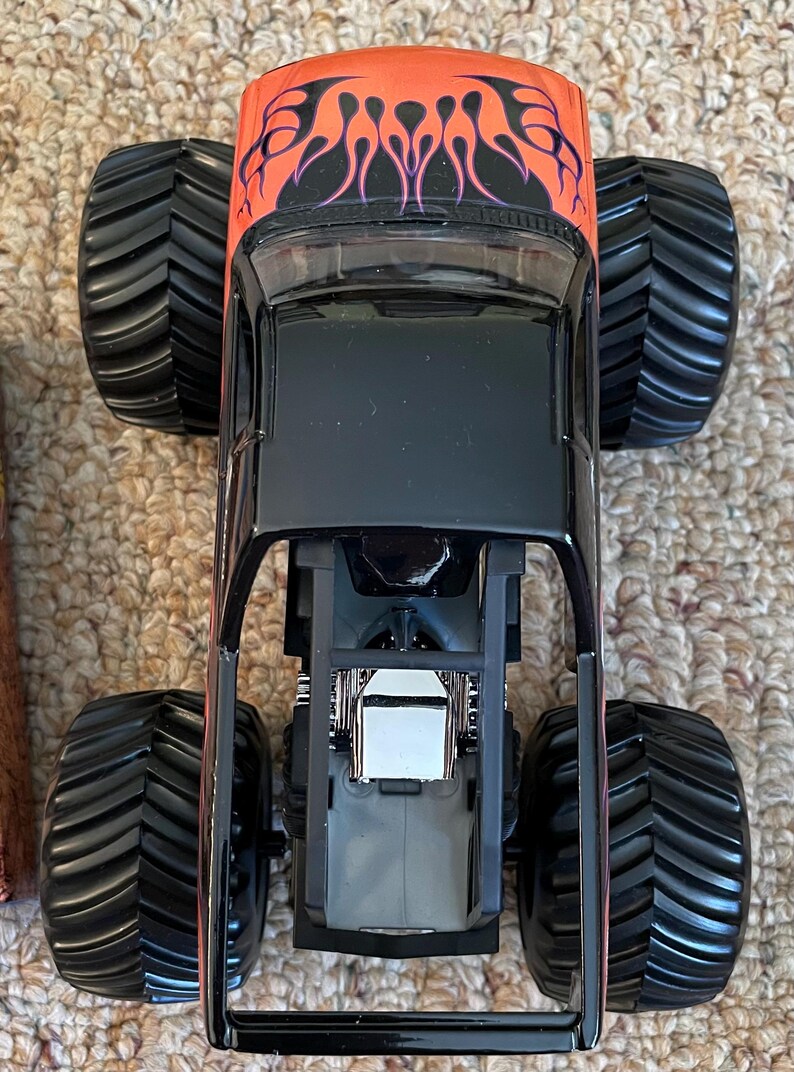 May include: A black monster truck toy with large black tires. The top of the truck is orange with a flame design. The truck's frame is black, and the engine is visible. The toy is designed for children.