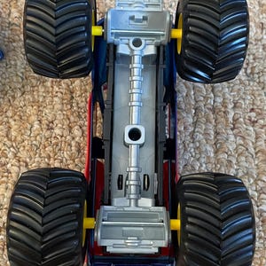 Op de afbeelding: Een speelgoed monstertruck met een rode, blauwe en zilveren carrosserie. De truck heeft vier grote zwarte banden met gele assen. De onderkant van de truck is zilver.