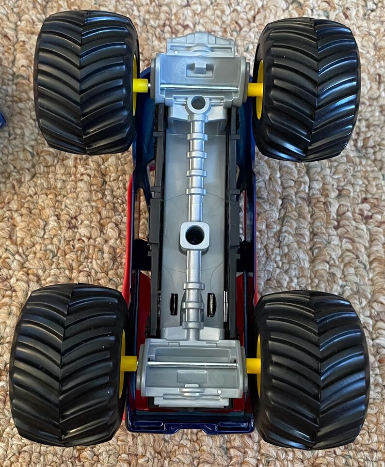 Op de afbeelding: Een speelgoed monstertruck met een rode, blauwe en zilveren carrosserie. De truck heeft vier grote zwarte banden met gele assen. De onderkant van de truck is zilver.