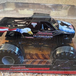 May include: Black monster truck toy with large tires, featuring an American flag design and an eagle graphic. The text "American Guardian" is displayed on the side. Packaged in a clear plastic container.