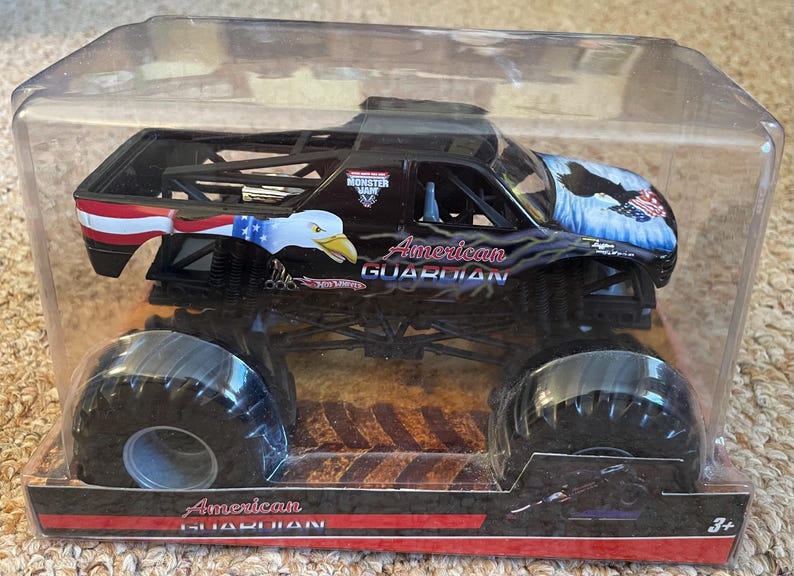 May include: Black monster truck toy with large tires, featuring an American flag design and an eagle graphic. The text "American Guardian" is displayed on the side. Packaged in a clear plastic container.