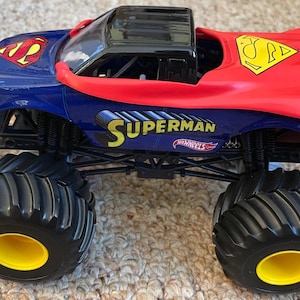 Op de afbeelding: Een Superman-thema Hot Wheels monstertruck speelgoed. De truck is voornamelijk blauw en rood, met het Superman-logo en het woord "Superman" in geel. Het heeft grote zwarte banden met gele velgen.
