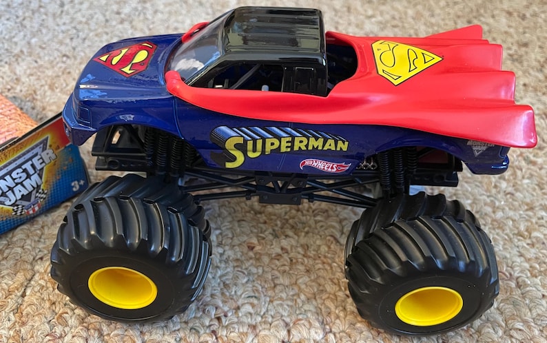 Op de afbeelding: Een Superman-thema Hot Wheels monstertruck speelgoed. De truck is voornamelijk blauw en rood, met het Superman-logo en het woord "Superman" in geel. Het heeft grote zwarte banden met gele velgen.