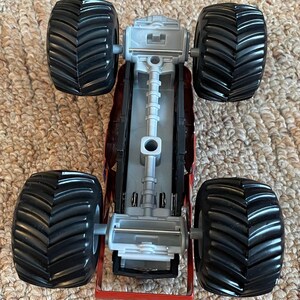 May include: A red and grey toy monster truck with four large black tyres. The truck has a detailed chassis and a red frame. The tyres have a deep tread pattern.