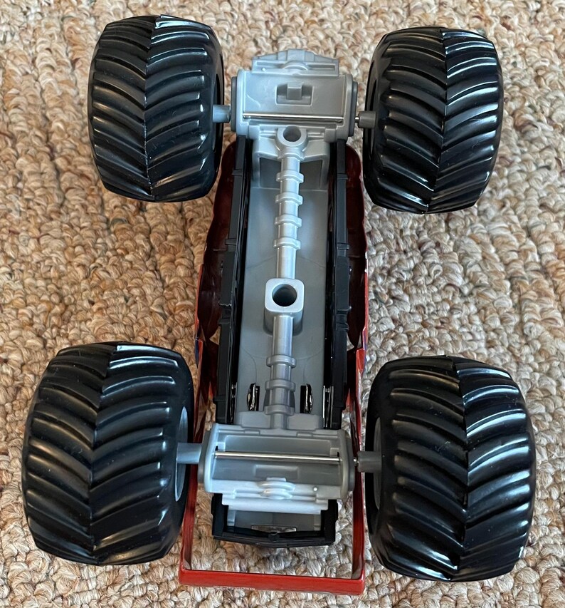 May include: A red and grey toy monster truck with four large black tyres. The truck has a detailed chassis and a red frame. The tyres have a deep tread pattern.