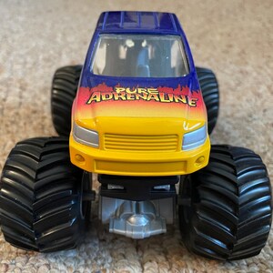 Może przedstawiać: Zabawkowy monster truck z dużymi czarnymi oponami. Kabina jest niebiesko-ż&oacute;łta z napisem "PURE ADRENALINE" w kolorze czerwonym i pomarańczowym. Ciężar&oacute;wka ma srebrne podwozie i jest przeznaczona do zabawy w terenie.