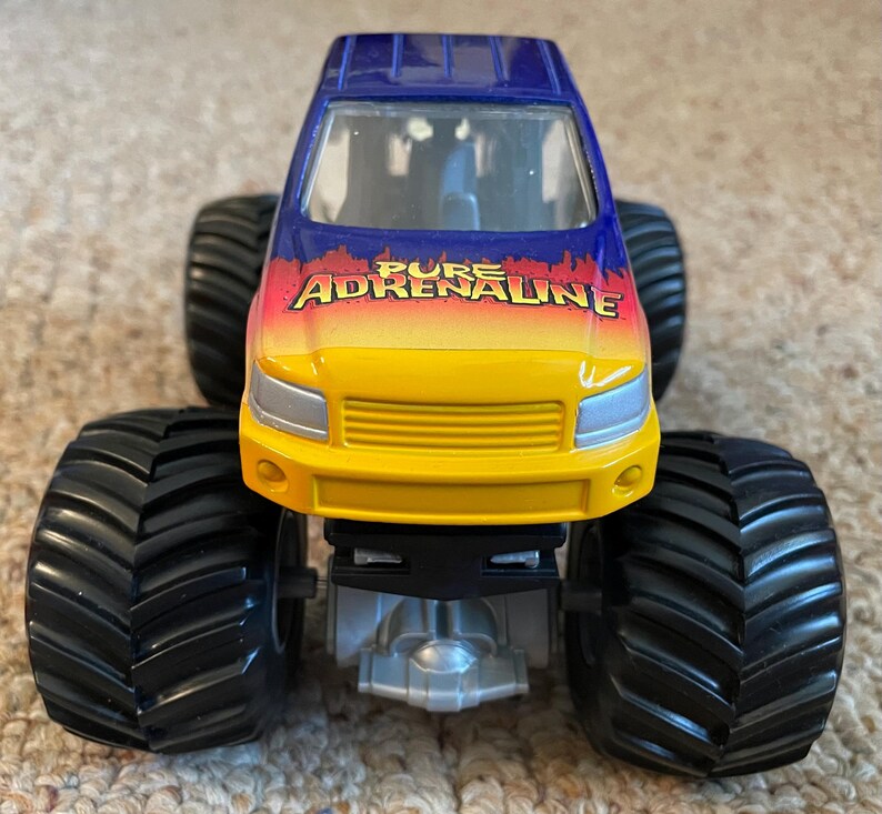 Może przedstawiać: Zabawkowy monster truck z dużymi czarnymi oponami. Kabina jest niebiesko-ż&oacute;łta z napisem "PURE ADRENALINE" w kolorze czerwonym i pomarańczowym. Ciężar&oacute;wka ma srebrne podwozie i jest przeznaczona do zabawy w terenie.