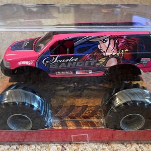 May include: A pink and black Hot Wheels monster truck toy, packaged in a clear plastic case. The truck features large black tires and the words "Scarlet Bandit" with an illustration of a woman on the side.