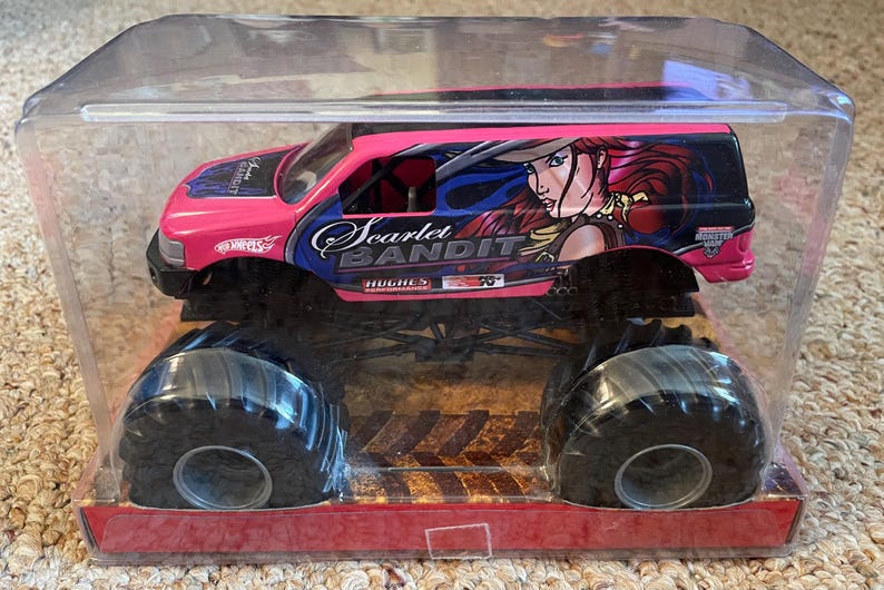 May include: A pink and black Hot Wheels monster truck toy, packaged in a clear plastic case. The truck features large black tires and the words "Scarlet Bandit" with an illustration of a woman on the side.