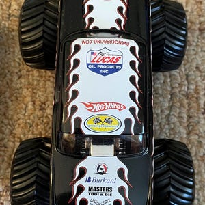 Puede incluir: Cami&oacute;n monstruo Hot Wheels negro con neum&aacute;ticos negros grandes. El cami&oacute;n presenta gr&aacute;ficos de llamas blancas y rojas y logotipos de Lucas Oil Products, Hot Wheels y Burkard Masters Tool & Die.