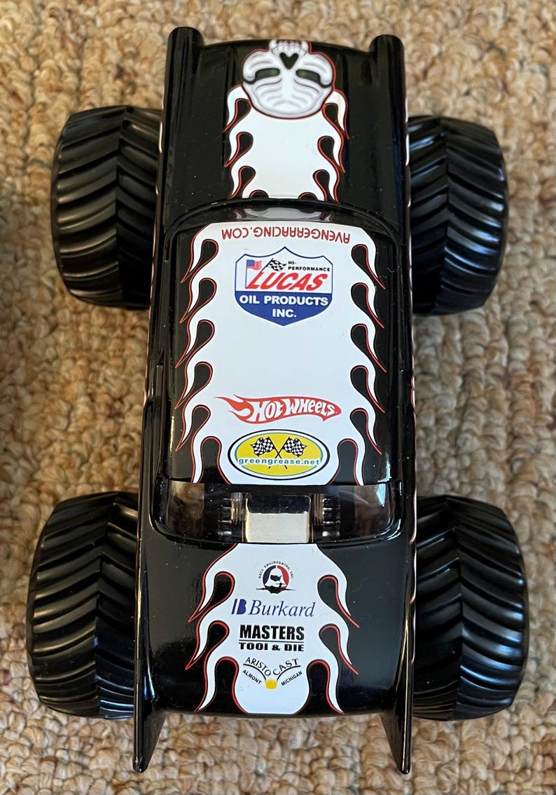 Puede incluir: Cami&oacute;n monstruo Hot Wheels negro con neum&aacute;ticos negros grandes. El cami&oacute;n presenta gr&aacute;ficos de llamas blancas y rojas y logotipos de Lucas Oil Products, Hot Wheels y Burkard Masters Tool & Die.