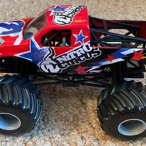 May include: A red and black toy monster truck with the words "Nitro Circus" and star graphics. The truck has large black tyres and a roll cage. The truck is a collectible toy.