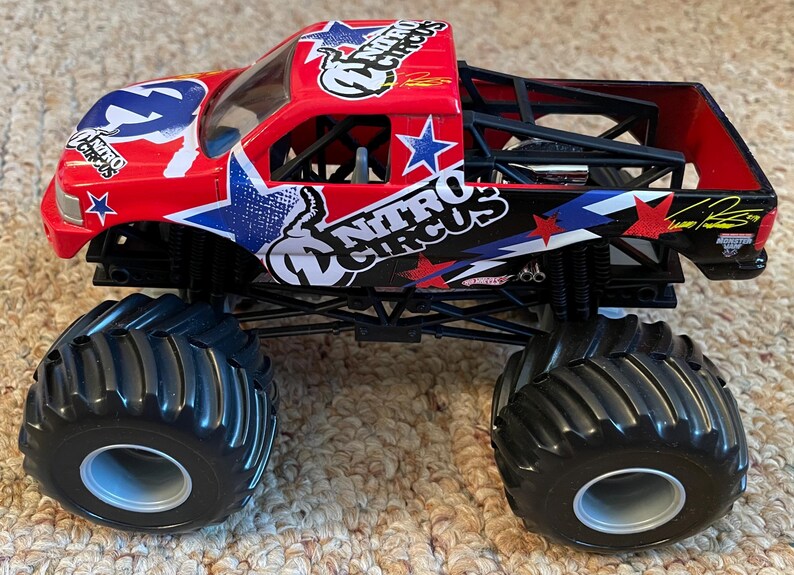 May include: A red and black toy monster truck with the words "Nitro Circus" and star graphics. The truck has large black tyres and a roll cage. The truck is a collectible toy.