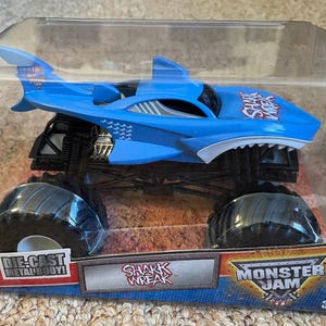 May include: A blue die-cast monster truck toy with a shark design, featuring large black tyres and the text "SHARK WREAK". The toy is packaged in a clear plastic container with "MONSTER JAM" branding.
