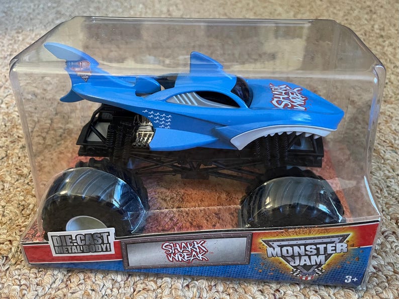 May include: A blue die-cast monster truck toy with a shark design, featuring large black tyres and the text "SHARK WREAK". The toy is packaged in a clear plastic container with "MONSTER JAM" branding.
