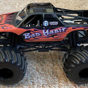 May include: A black and orange toy monster truck with large black tires. The truck has the words "Bad Habit" in purple and white, with flame graphics. The truck also has the Hot Wheels Monster Jam logo.