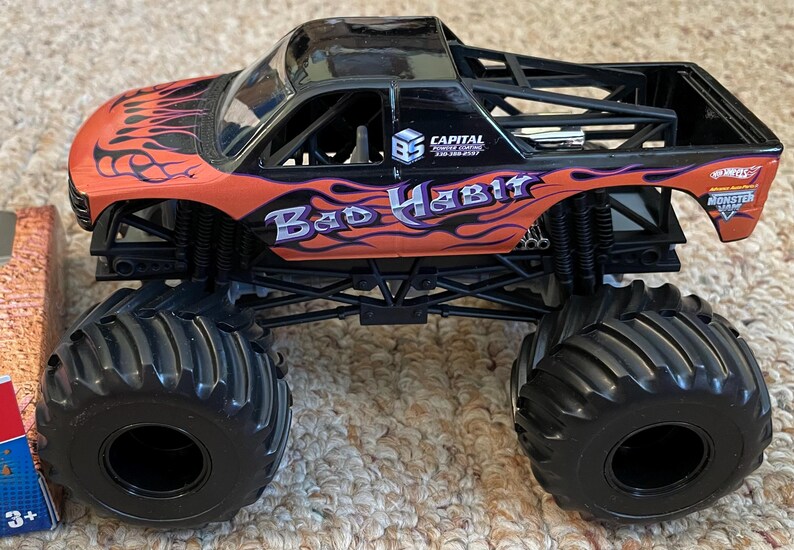 May include: A black and orange toy monster truck with large black tires. The truck has the words "Bad Habit" in purple and white, with flame graphics. The truck also has the Hot Wheels Monster Jam logo.
