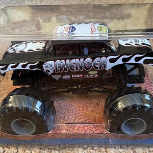 Puede incluir: Un cami&oacute;n monstruo de juguete en blanco y negro llamado "Avenger" en una caja de pl&aacute;stico transparente. El cami&oacute;n tiene neum&aacute;ticos negros grandes y un dise&ntilde;o de llamas. La cabina presenta el logotipo de Hot Wheels.