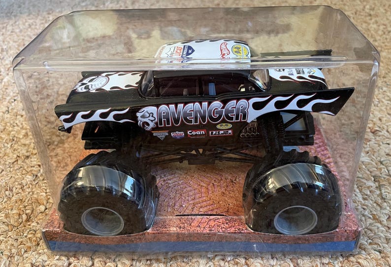 Puede incluir: Un cami&oacute;n monstruo de juguete en blanco y negro llamado "Avenger" en una caja de pl&aacute;stico transparente. El cami&oacute;n tiene neum&aacute;ticos negros grandes y un dise&ntilde;o de llamas. La cabina presenta el logotipo de Hot Wheels.