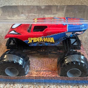 May include: A Spider-Man monster truck toy in a clear plastic package. The truck is red and blue with the Spider-Man logo and design. The large black tires are visible. The packaging sits on a brown and blue base.