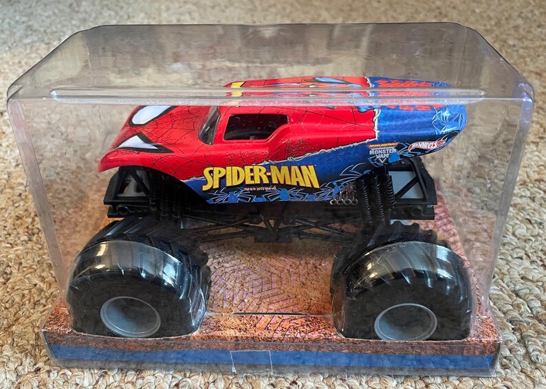 May include: A Spider-Man monster truck toy in a clear plastic package. The truck is red and blue with the Spider-Man logo and design. The large black tires are visible. The packaging sits on a brown and blue base.