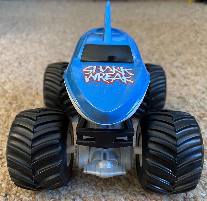 May include: A blue and white toy monster truck with large black tyres. The truck has a shark fin on top and the words "SHARK WREAK" in red and white on the hood.