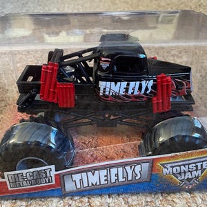 May include: Black and red die-cast metal body monster truck toy in a clear plastic package. The truck has large black tires and the words "TIME FLYS" on the side. The package also features the Monster Jam logo.