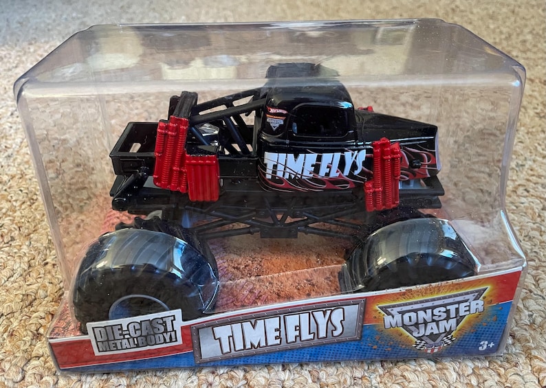 May include: Black and red die-cast metal body monster truck toy in a clear plastic package. The truck has large black tires and the words "TIME FLYS" on the side. The package also features the Monster Jam logo.