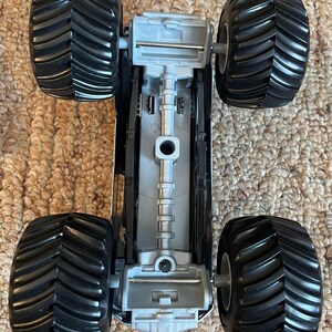 Pu&ograve; includere: Un giocattolo di monster truck grigio e nero con grandi pneumatici neri e strutturati. Il lato inferiore del camion &egrave; visibile, mostrando il telaio e i dettagli delle sospensioni. Il giocattolo &egrave; progettato per il gioco fuoristrada.