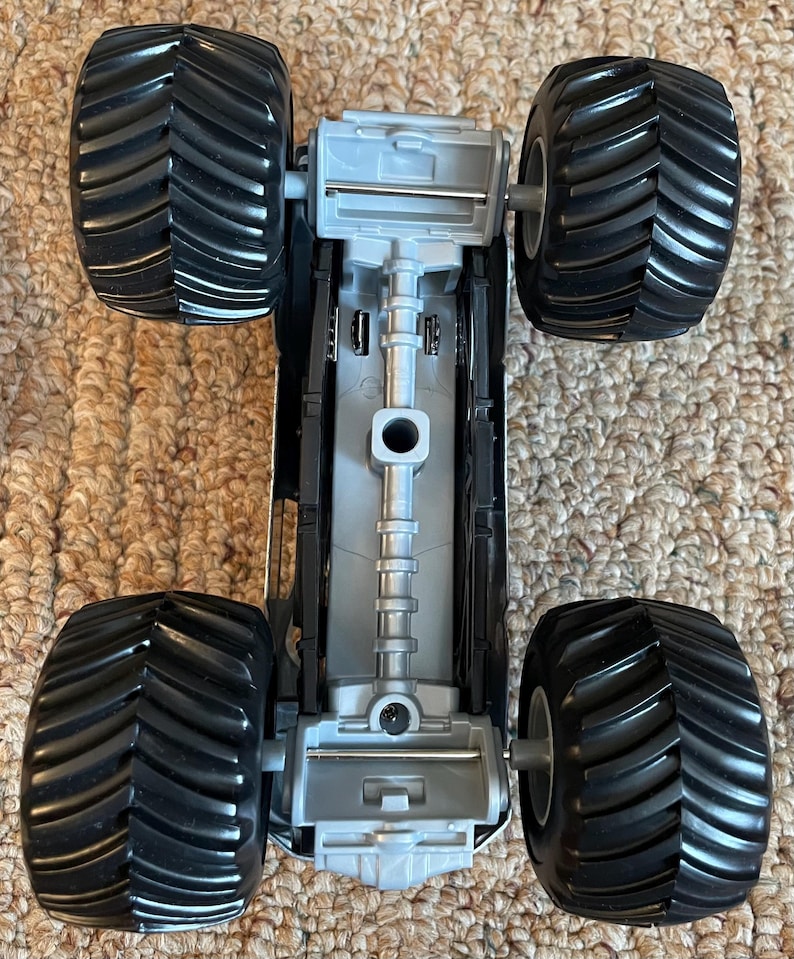 Pu&ograve; includere: Un giocattolo di monster truck grigio e nero con grandi pneumatici neri e strutturati. Il lato inferiore del camion &egrave; visibile, mostrando il telaio e i dettagli delle sospensioni. Il giocattolo &egrave; progettato per il gioco fuoristrada.