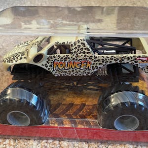 May include: A toy monster truck in a clear plastic package. The truck has a cream-colored body with a leopard print design and the word "Pouncer" in orange. It has large black tires and is displayed on a red base.
