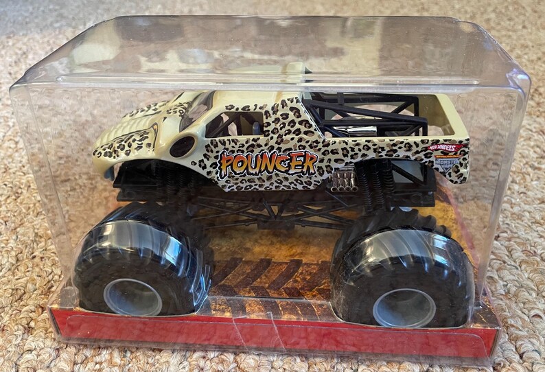 May include: A toy monster truck in a clear plastic package. The truck has a cream-colored body with a leopard print design and the word "Pouncer" in orange. It has large black tires and is displayed on a red base.