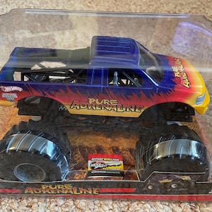 Może przedstawiać: Zabawka ciężar&oacute;wki potwora Hot Wheels w przezroczystym plastikowym opakowaniu. Ciężar&oacute;wka jest niebieska, ż&oacute;łta i czerwona z napisem "Pure Adrenaline". Ma duże czarne opony i szczeg&oacute;łowe podwozie. Opakowanie zawiera dodatkowy tekst i grafikę.