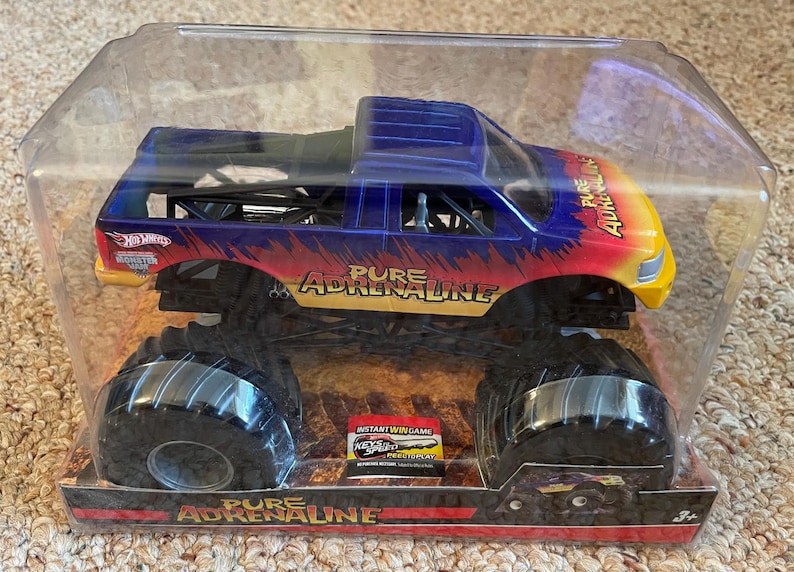 Może przedstawiać: Zabawka ciężar&oacute;wki potwora Hot Wheels w przezroczystym plastikowym opakowaniu. Ciężar&oacute;wka jest niebieska, ż&oacute;łta i czerwona z napisem "Pure Adrenaline". Ma duże czarne opony i szczeg&oacute;łowe podwozie. Opakowanie zawiera dodatkowy tekst i grafikę.