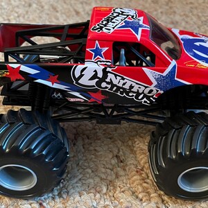 May include: A red and black toy monster truck with large black tyres. The truck features the "Nitro Circus" logo with stars and stripes. The truck has a black roll cage and is sitting on a beige carpet.
