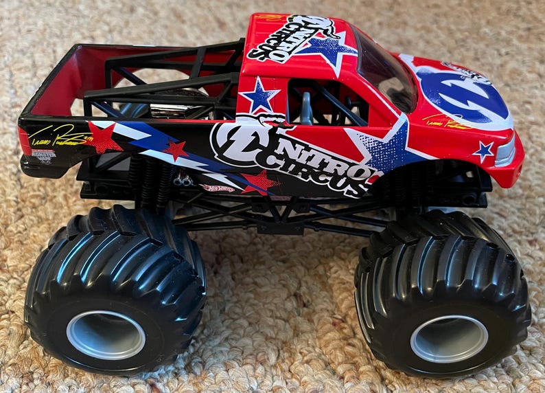 May include: A red and black toy monster truck with large black tyres. The truck features the "Nitro Circus" logo with stars and stripes. The truck has a black roll cage and is sitting on a beige carpet.
