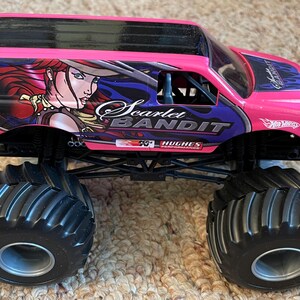May include: A pink and black toy monster truck with large black tires. The truck features the words "Scarlet Bandit" and a graphic of a woman with red hair. The Hot Wheels logo is visible on the side.