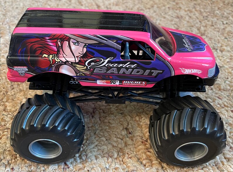 May include: A pink and black toy monster truck with large black tires. The truck features the words "Scarlet Bandit" and a graphic of a woman with red hair. The Hot Wheels logo is visible on the side.